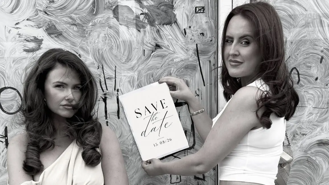 Lauren and Nicole holding a save-the-date card reading 15.08.26 against a painted-out salon window